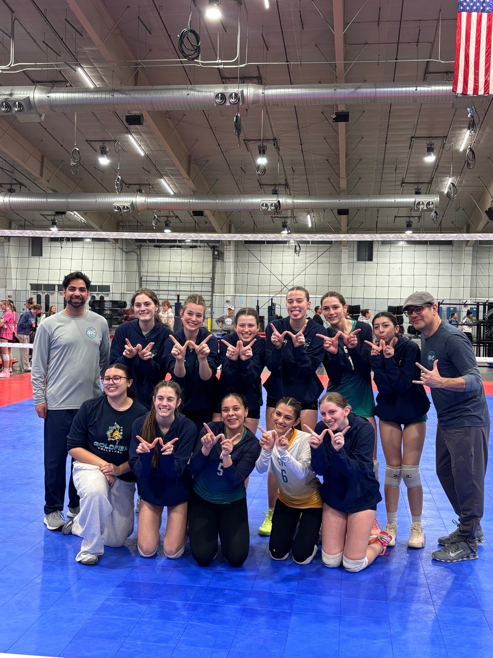 A large group of volleyball players and two coaches posing together on the court, with some players making 'W' sign gestures with their hands.